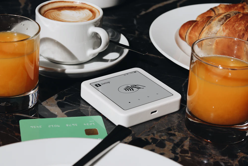 A small contactless card reader sitting on a café table between a cappuccino, a glass of orange juice and a croissant.