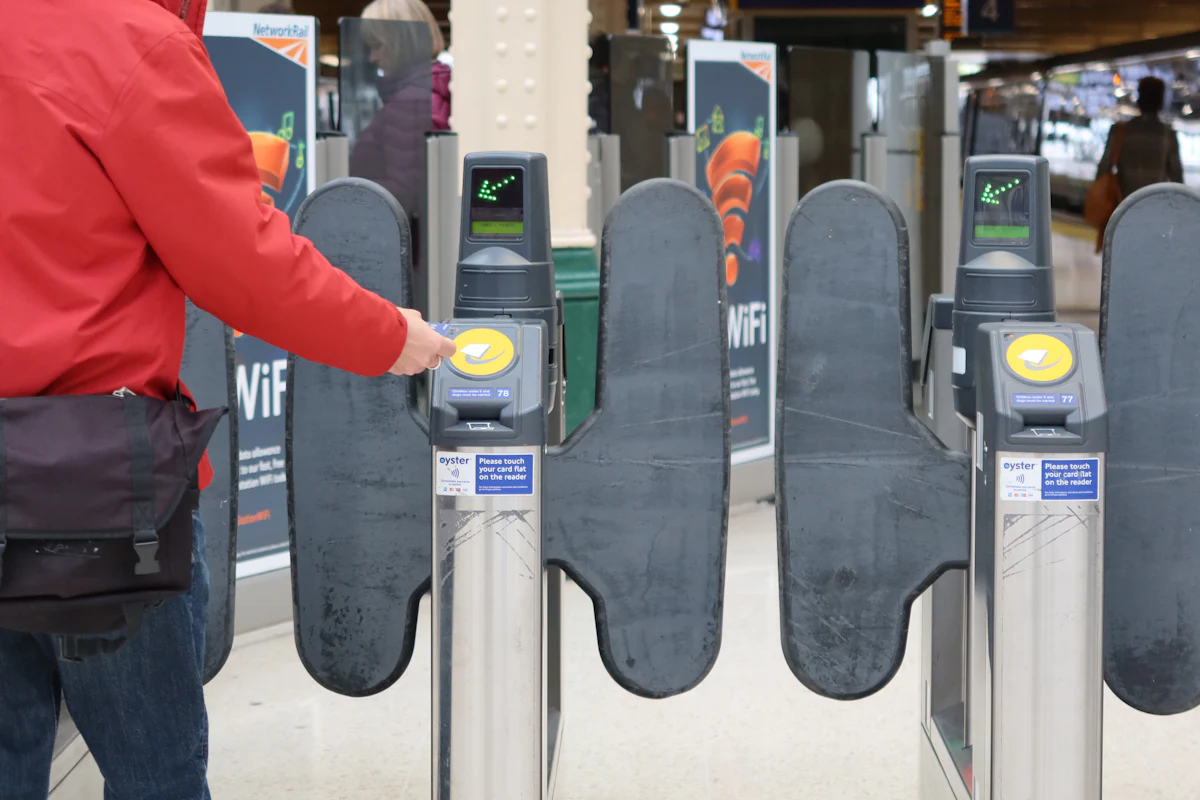 A commuter in a red jacket touches a travel card to the yellow reader on a railway station ticket gate.